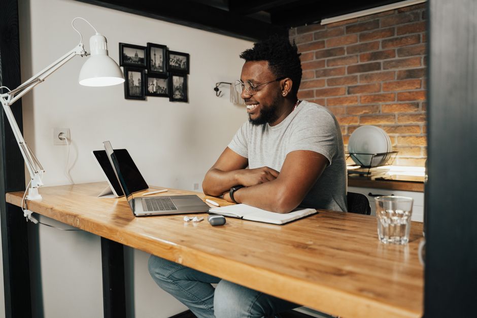 A man smiling at a standing desk with a laptop and a notebook open in front of him.