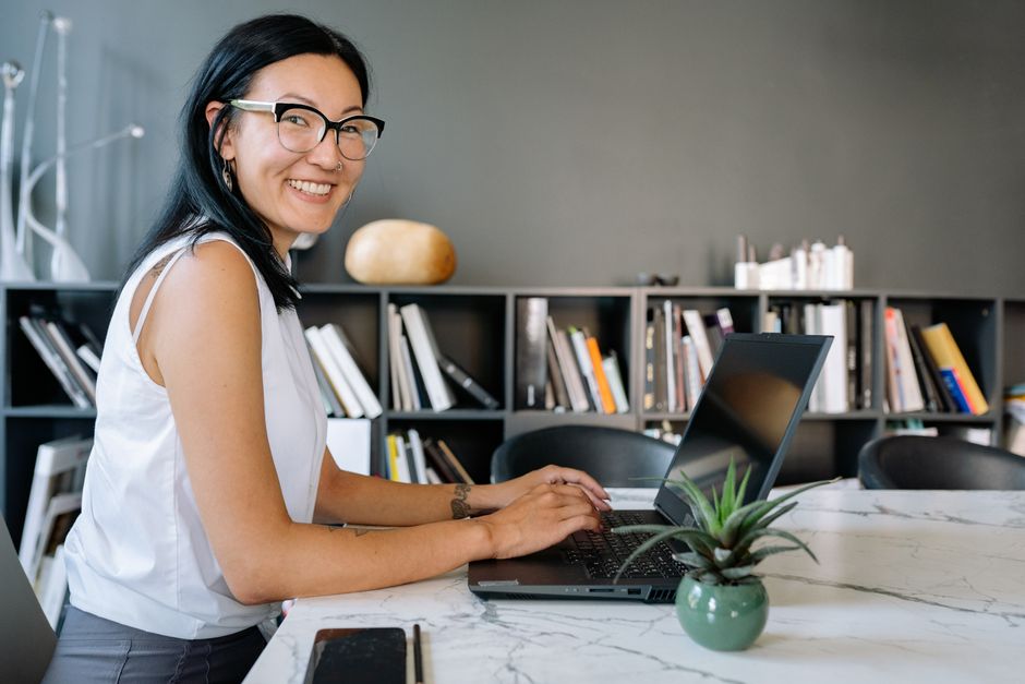A woman smiling as she works at a laptop beside a bookshelf.