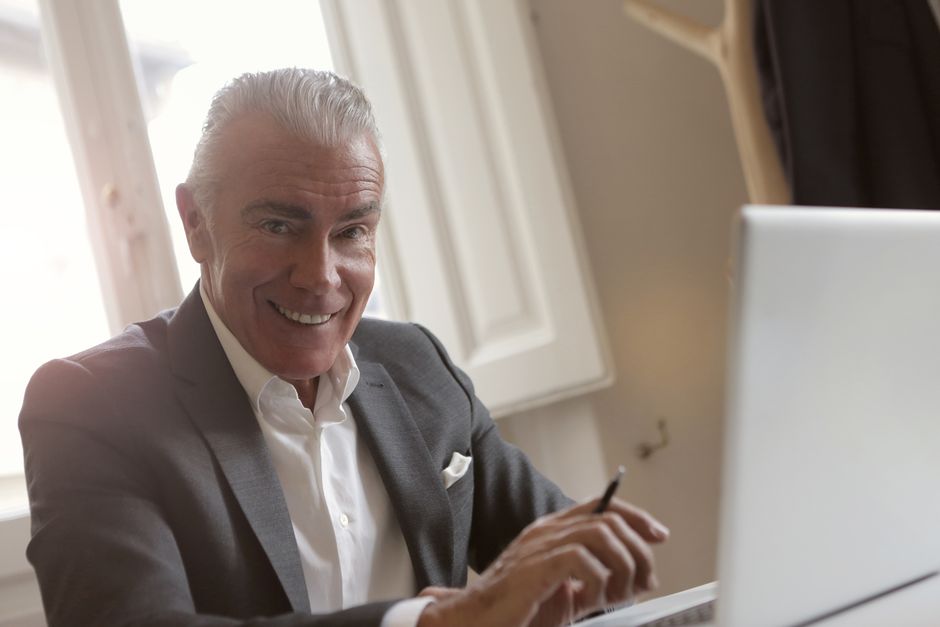 A grey-haired man smiling while working at a laptop, pen in hand.