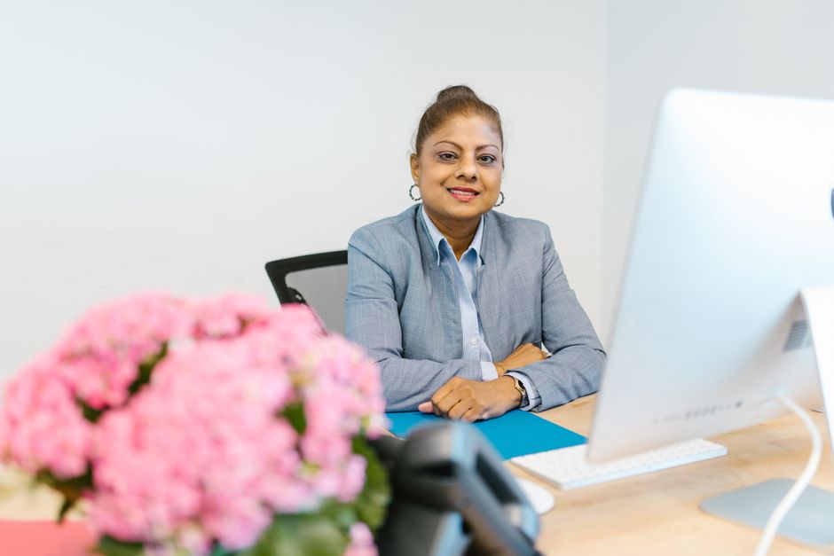 A woman in a blue suit smiling at her desk beside a computer.