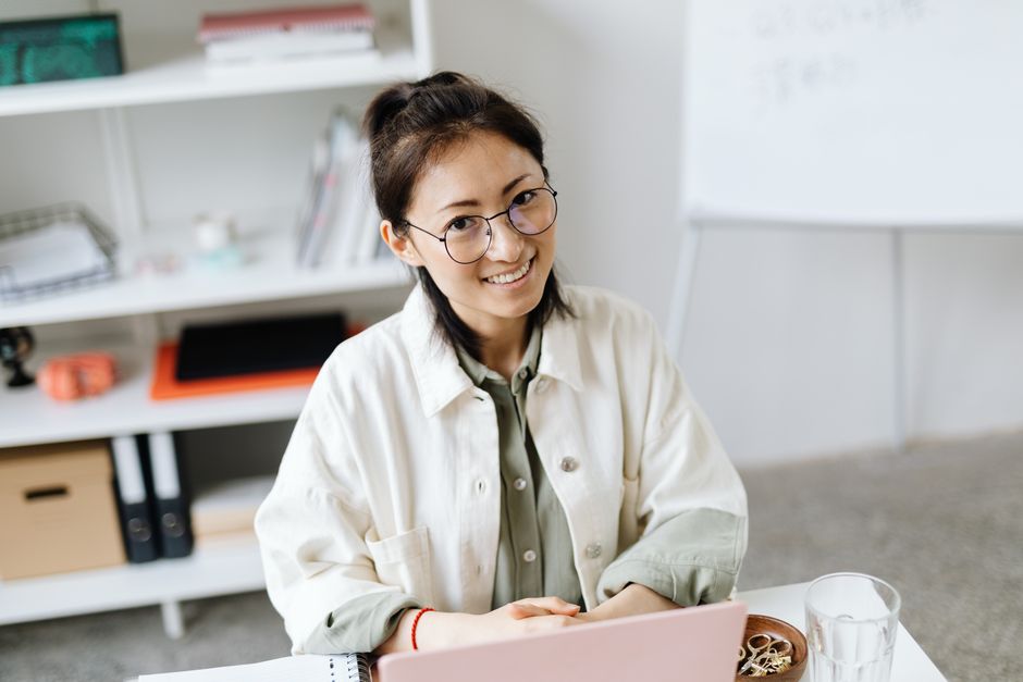 A woman in glasses smiling while working at a laptop in a bright office.